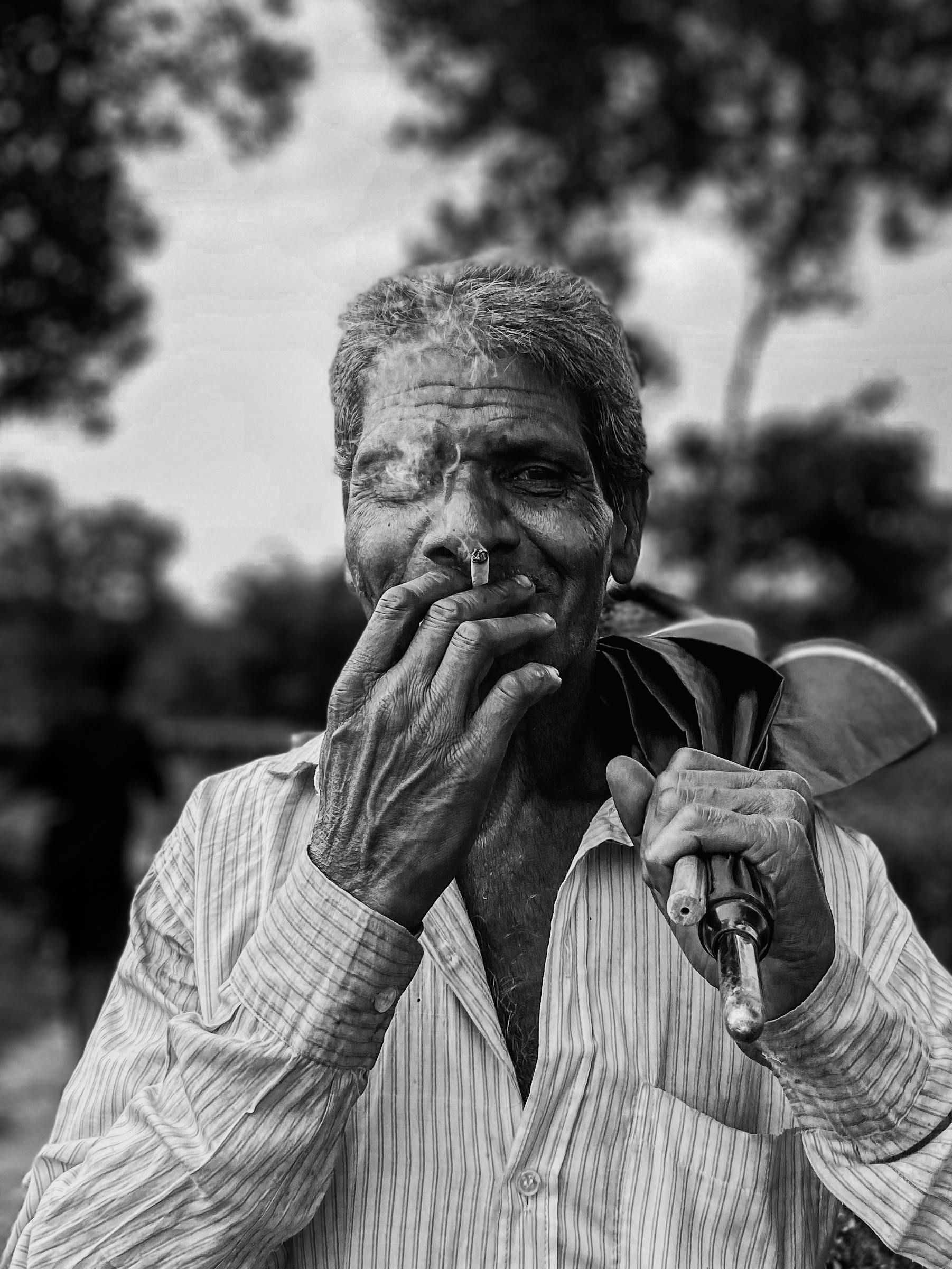 Man with weathered face holding a cigarette, wearing a striped shirt. Blurred outdoor background.