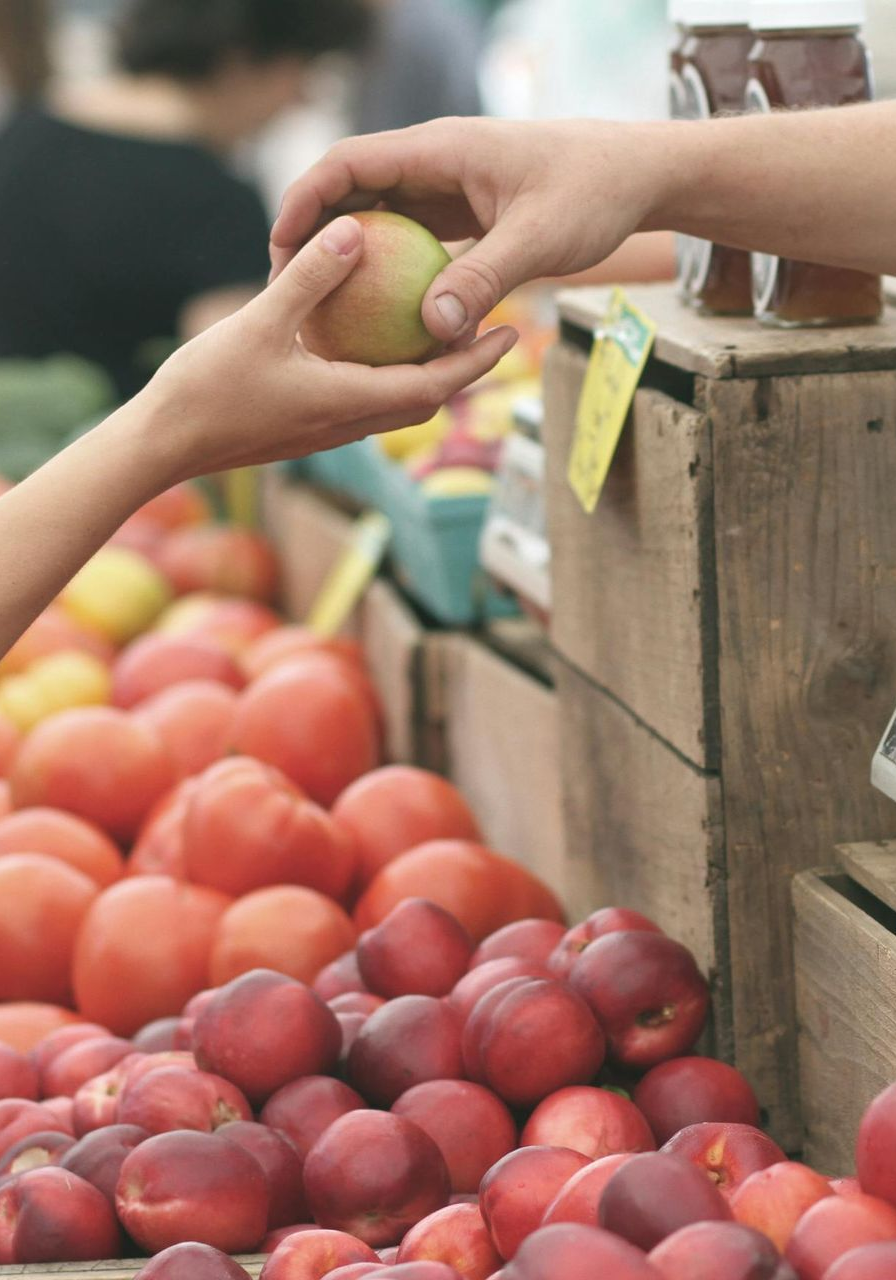 A hand passing a piece of fruit to another hand at a farmer's market, with produce displayed in front.