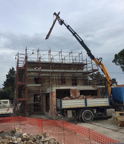 Cantiere edile con una gru che solleva materiali accanto a un edificio in mattoni impalcato e a un camion parcheggiato.