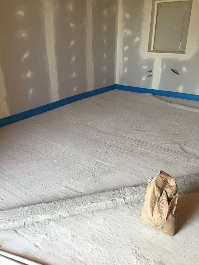 Stanza vuota con pareti in cartongesso non rifinite, nastro adesivo blu da imbianchino lungo i battiscopa e un sacchetto di carta sul pavimento.