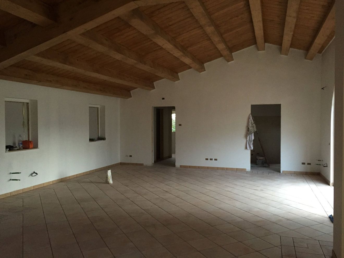 Stanza vuota e non rifinita con soffitto a travi in ​​legno, pavimento piastrellato e porte aperte.