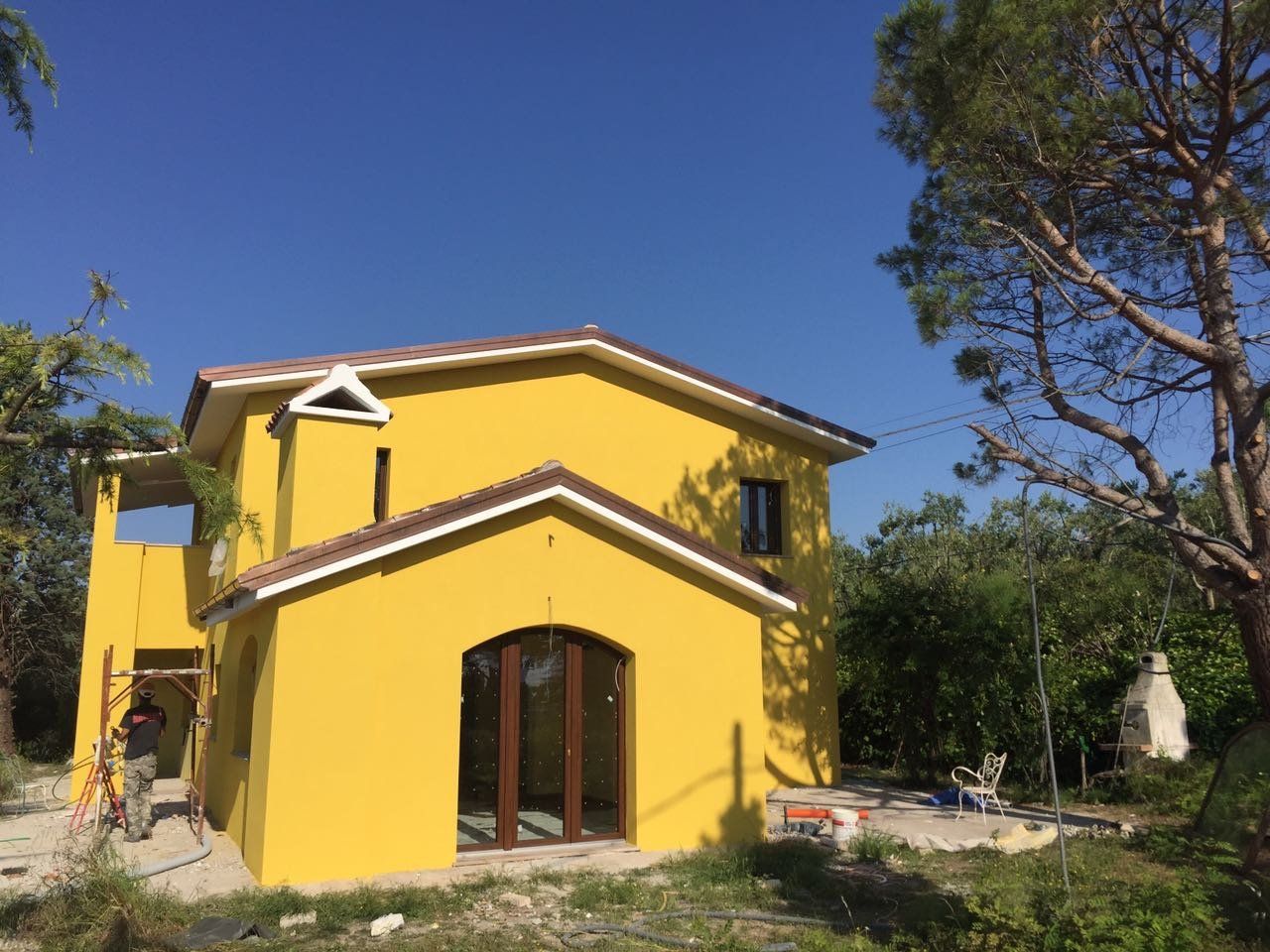 Casa gialla a due piani con finestre incorniciate di marrone e tetto a due falde sotto un cielo azzurro limpido.