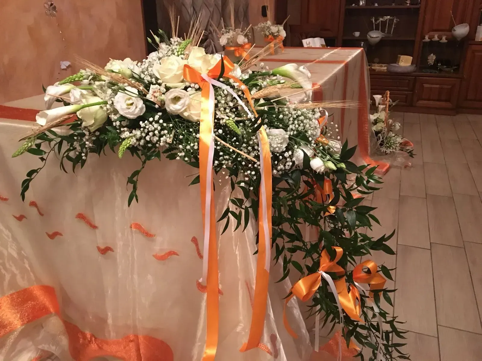 Composizione floreale bianca con nastri arancioni drappeggiata su un tavolo coperto da una tovaglia in una chiesa o in una sala ricevimenti.
