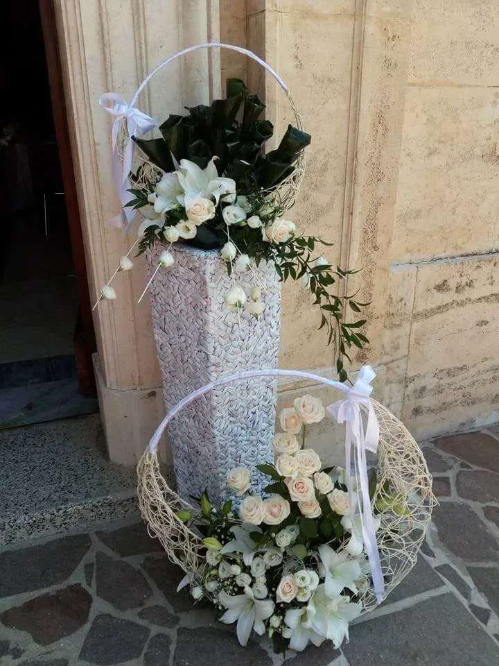 Due composizioni floreali bianche in cesti di vimini su un pavimento di pietra, decorate con nastri bianchi.