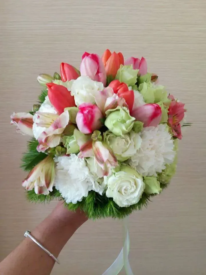 Bouquet da tenere in mano, composto da fiori bianchi, rosa e color corallo con verde e un nastro bianco su sfondo neutro.