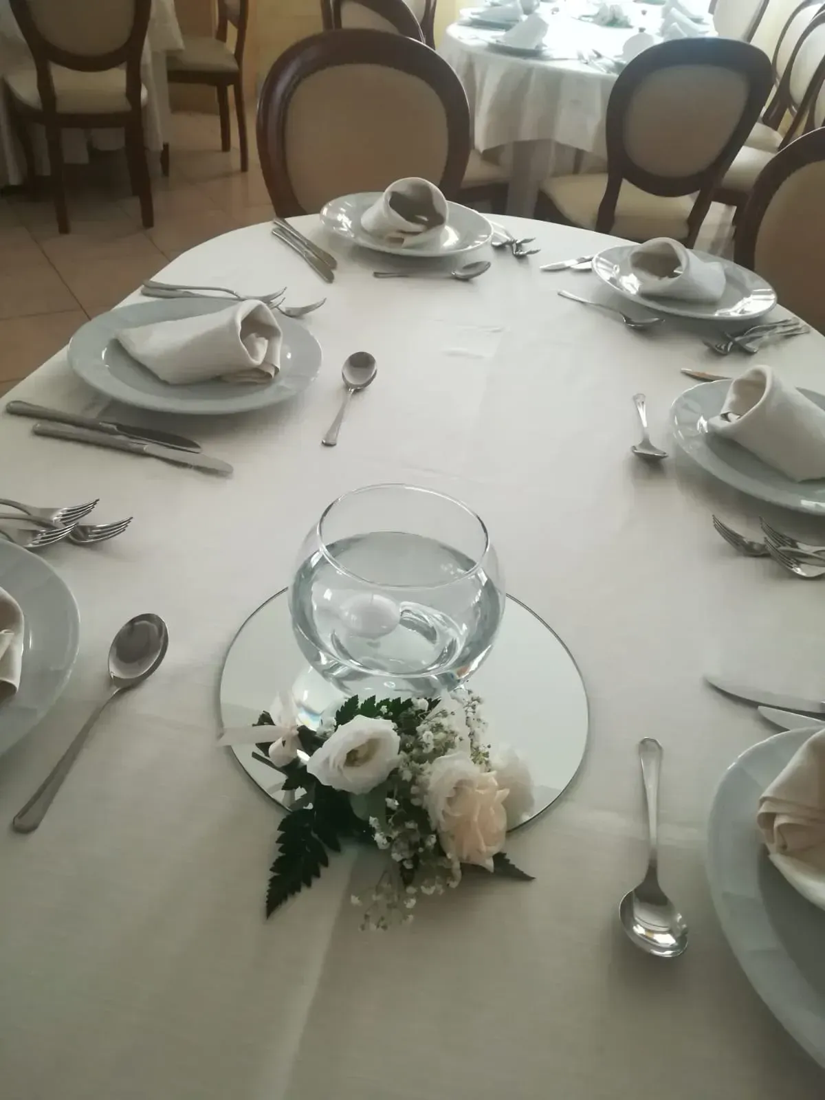 Tavolo rotondo per banchetto apparecchiato con piatti bianchi, posate, bicchieri e un centrotavola floreale su una tovaglia bianca.