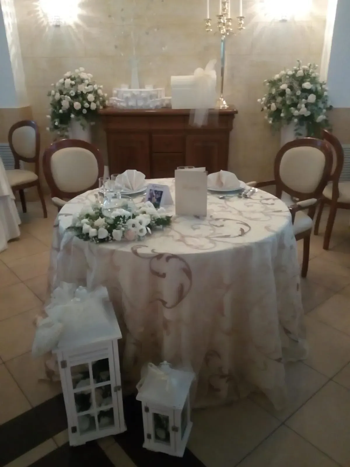 Elegante tavolo per il ricevimento di nozze con decorazioni floreali bianche, lanterne e sedie accanto a un caminetto in legno e candele.