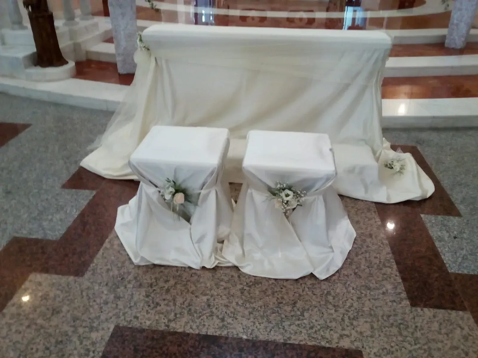 Panca drappeggiata di bianco con due sgabelli abbinati e decorazioni floreali nella navata di una chiesa.