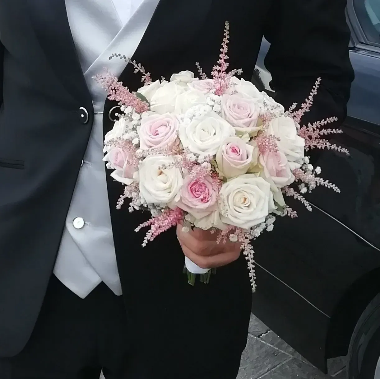 Persona in abito scuro che tiene in mano un mazzo di rose bianche e rosa pallido con fiori di riempimento rosa.