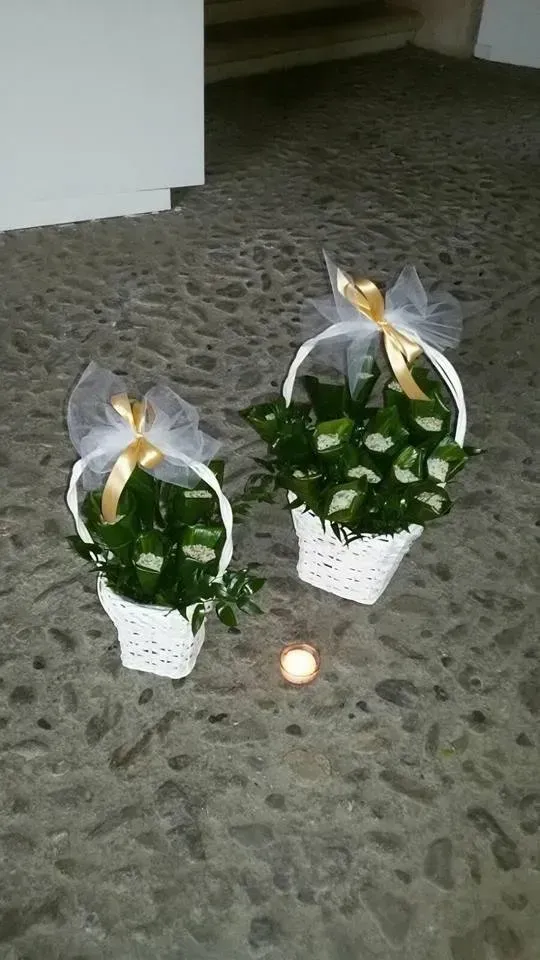 Due piccoli cestini regalo bianchi con fogliame verde e fiocchi di nastro su un pavimento grigio, con una candela in mezzo.