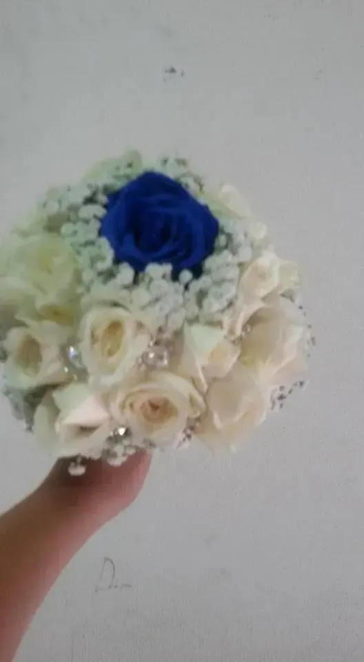 Piccolo mazzo di rose color crema con un fiore blu, tenuto in mano su uno sfondo neutro.