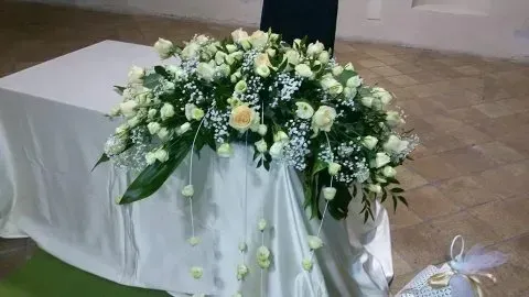 Composizione floreale bianca su un tavolo drappeggiato durante un evento al chiuso, con foglie verdi e piccoli fiori pendenti.