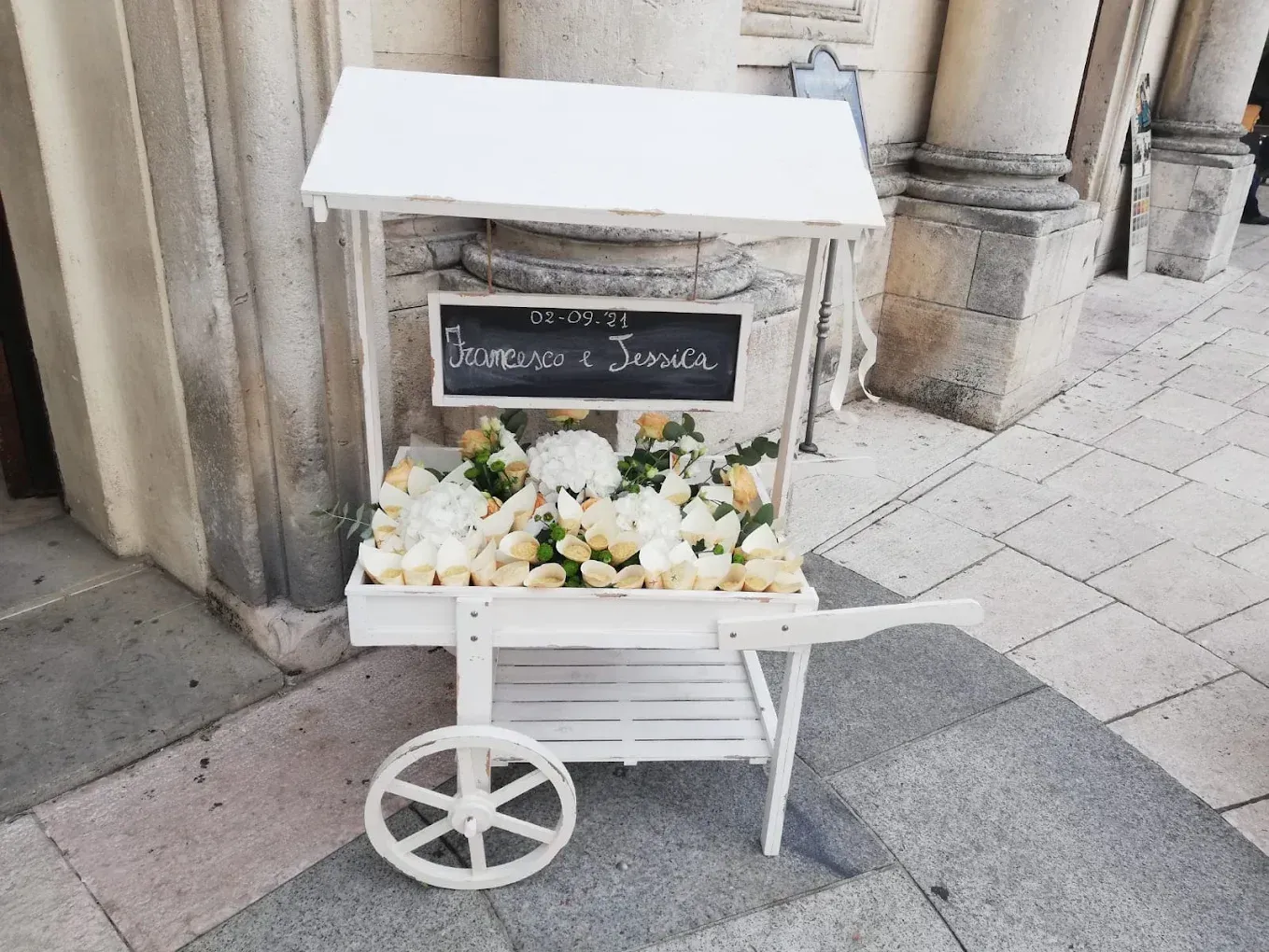 Carretto di fiori bianco accanto a un muro di pietra con una lavagna su cui è scritto