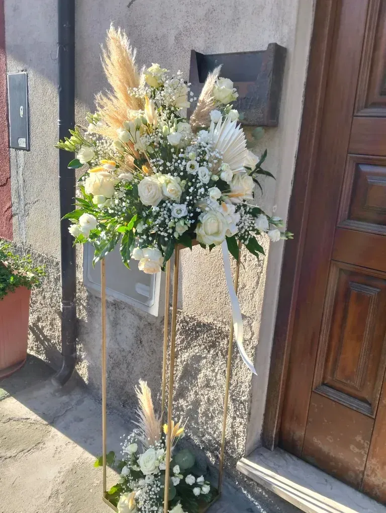 Composizione floreale bianca in piedi su cavalletti dorati accanto a una porta di legno.