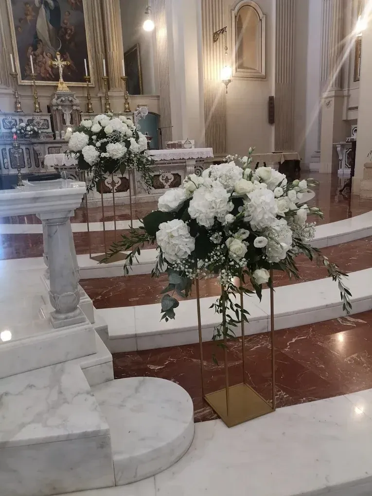 Composizioni floreali bianche su supporti dorati decorano l'altare di una chiesa con gradini in marmo e pavimento in piastrelle rosse.
