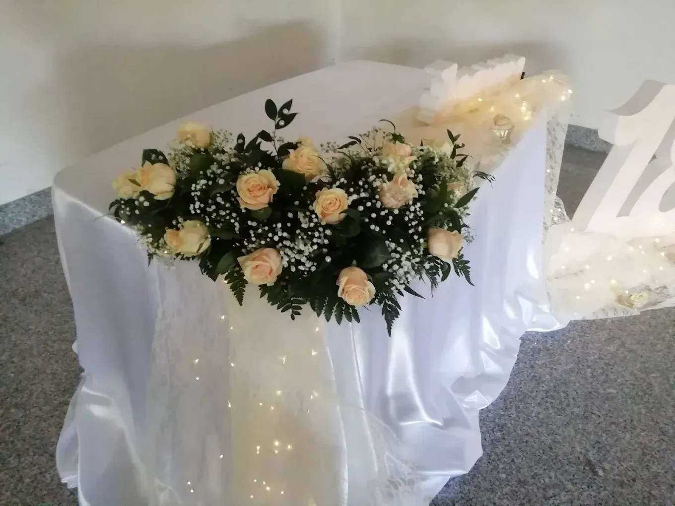Tavolo nuziale drappeggiato di bianco con centrotavola floreale color pesca e bianco e lucine scintillanti.