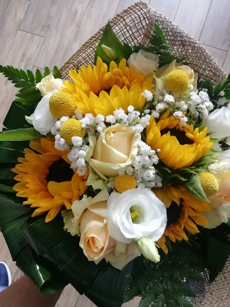 Bouquet di girasoli, rose bianche e fiori gialli avvolti in foglie verdi e rete su un pavimento piastrellato.