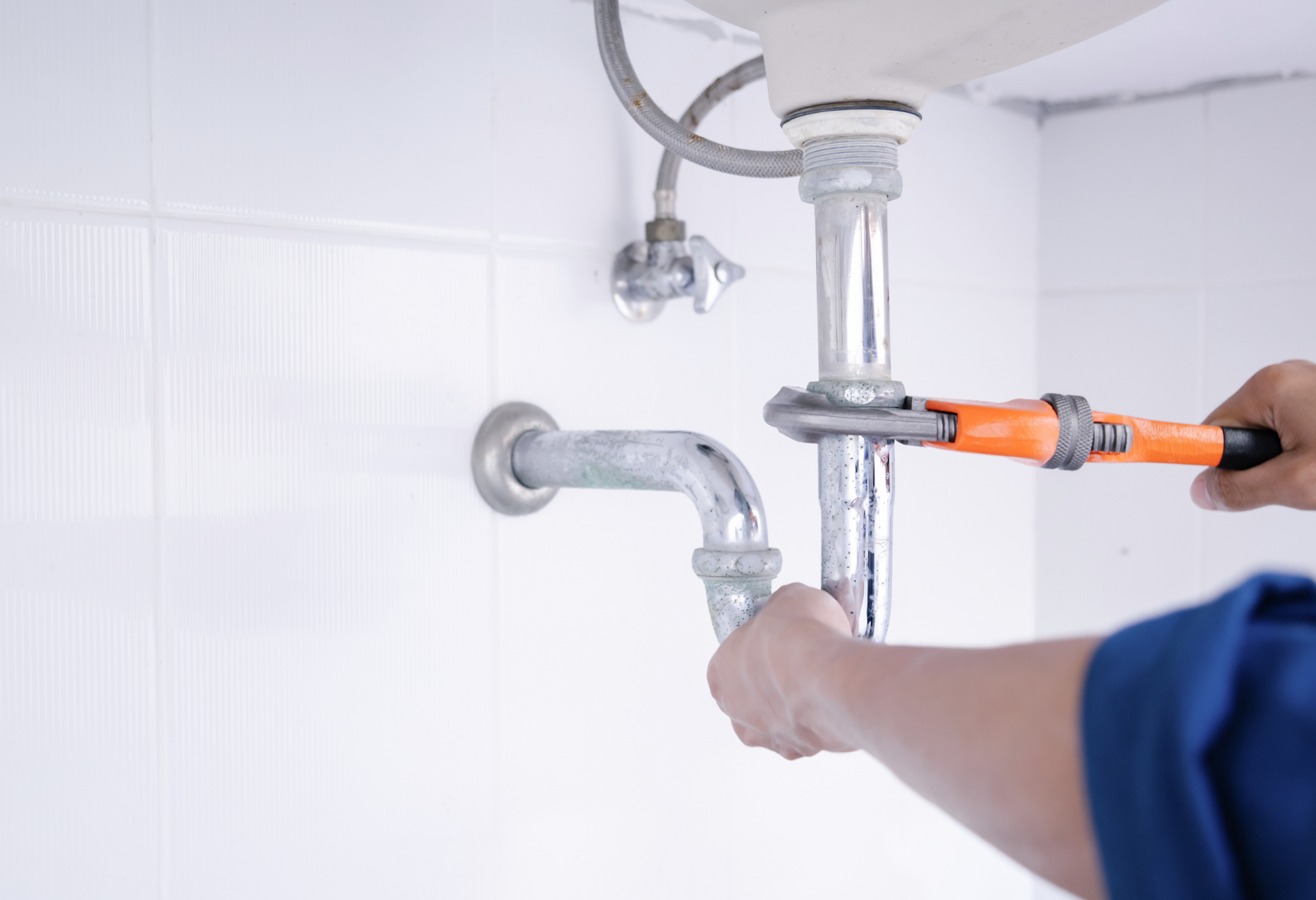 Plumber using a wrench on chrome pipes under a sink in a white-tiled room.