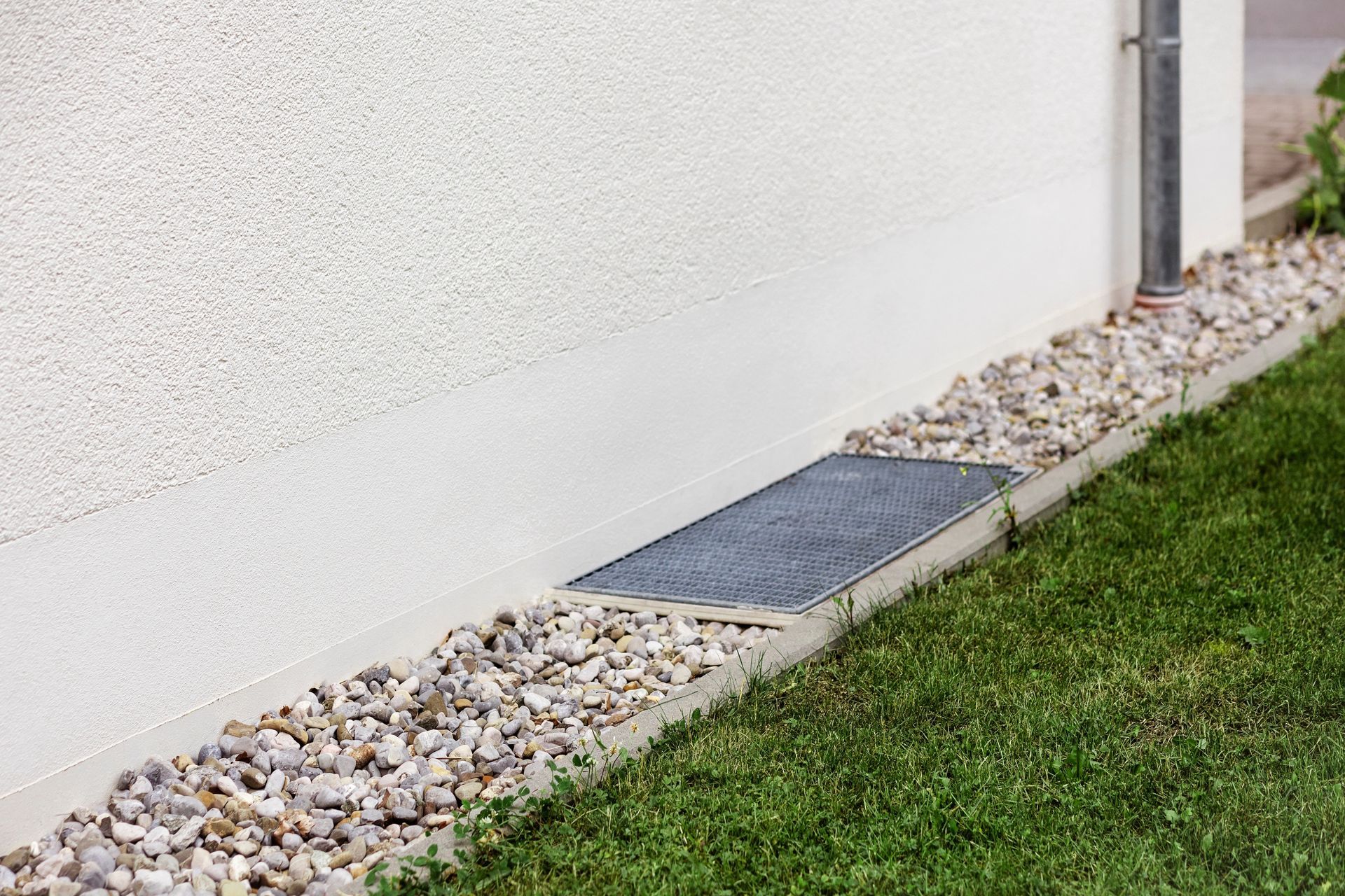 Basement window well beside a white stucco wall, edged with gravel and grass.