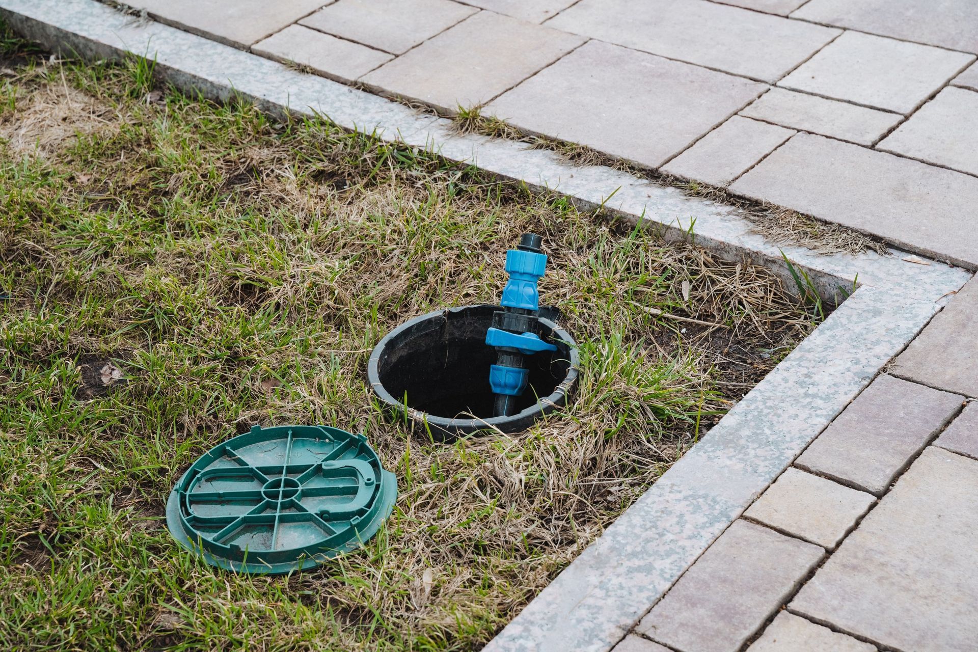 Open utility box in a paved walkway, with a blue pipe visible and the green lid beside it