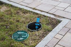 Open water meter box with blue valve beside a paved walkway and grass border