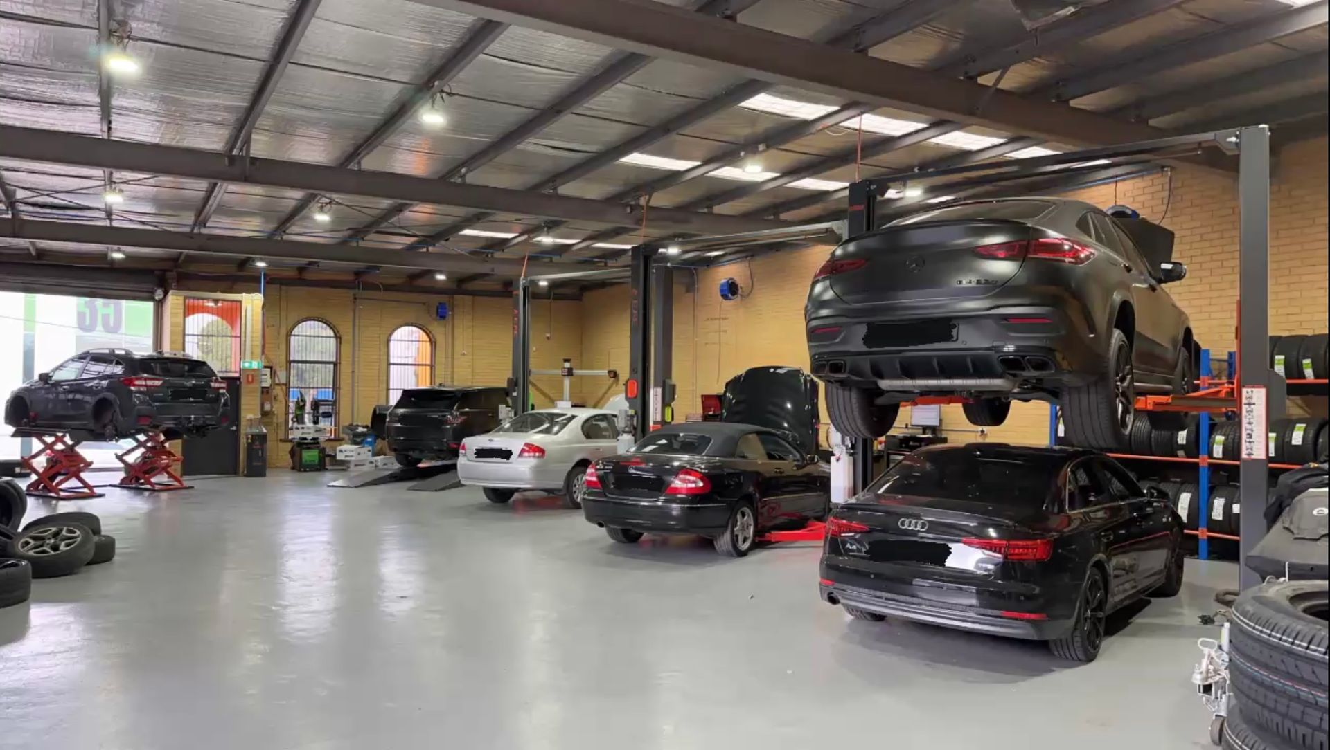 Cars on lifts and floor in a vehicle repair shop. Tires stacked on a shelf.