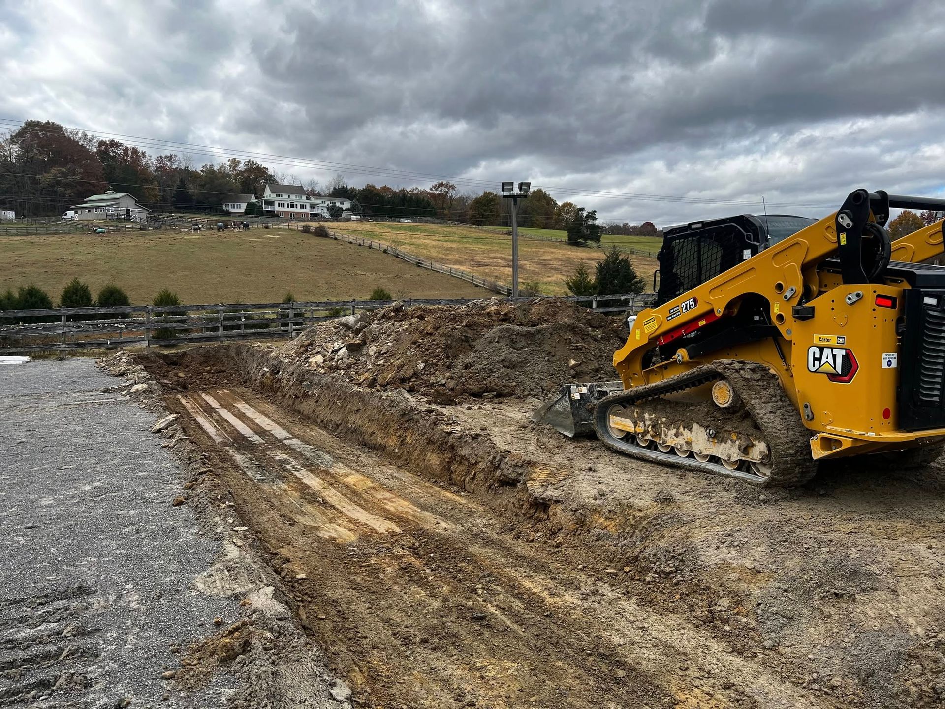 Construction site with a yellow excavator on a dirt path near bare trees and concrete foundations.