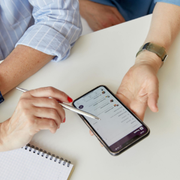 Hands using a stylus on a smartphone over a notebook on a white table