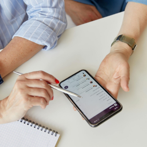 Hands using a stylus on a smartphone over a notebook on a white table