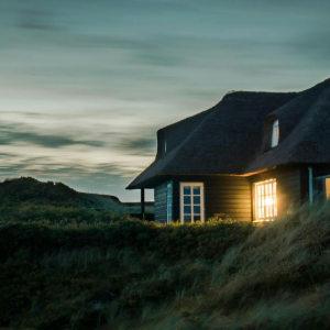 Cozy cottage with glowing window at dusk on a grassy hillside under a cloudy sky