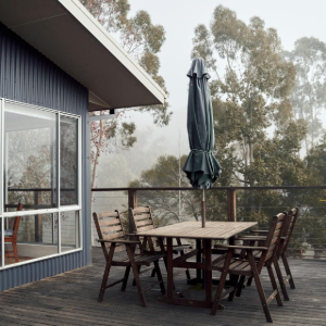 Outdoor deck with wooden table, chairs, and closed umbrella beside a modern house, overlooking trees and fog.