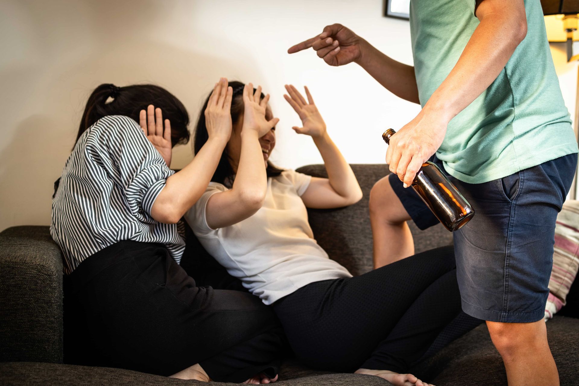 Two people on couch react in fear as another, holding a bottle, points at them.