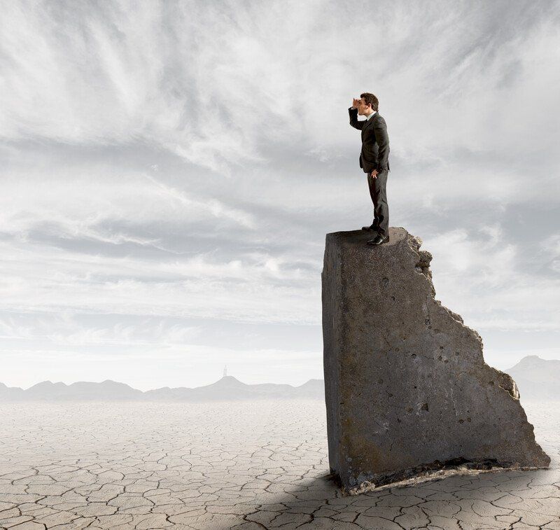 Man in suit atop crumbling pillar, looking out over cracked desert landscape.