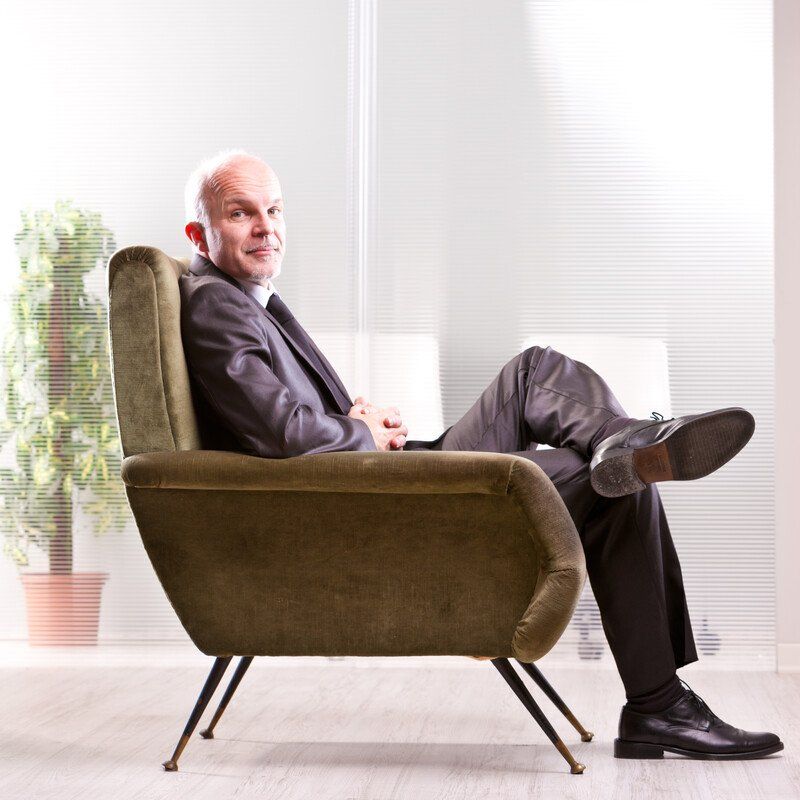 Bald man in a suit sits in an olive green chair, leg crossed, in an office with a plant.