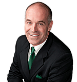 Bald man in a black suit with a green tie and pocket square, smiling.