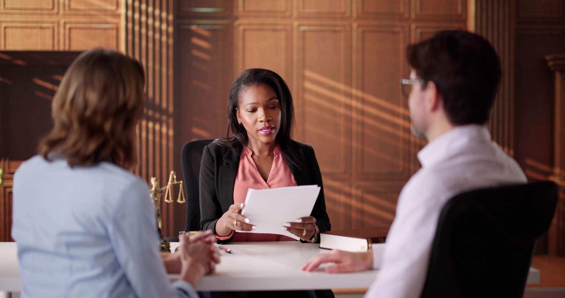 A woman in a black suit presents a document to a couple seated at a table in a wood-paneled office.