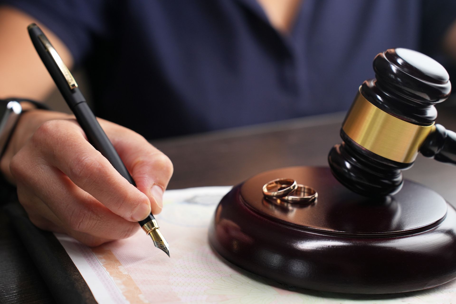 Close-up of a person signing a divorce document next to a gavel and wedding rings. Hand writing with pen near gavel and wedding rings on a wooden base.