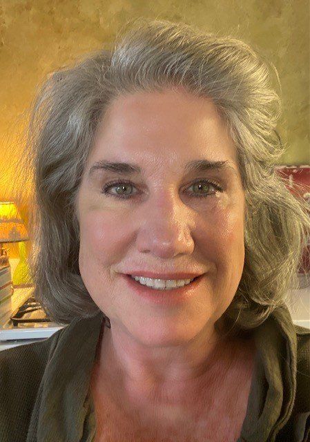 Woman with gray hair smiles, wearing an olive green shirt. Bright, indoor setting.