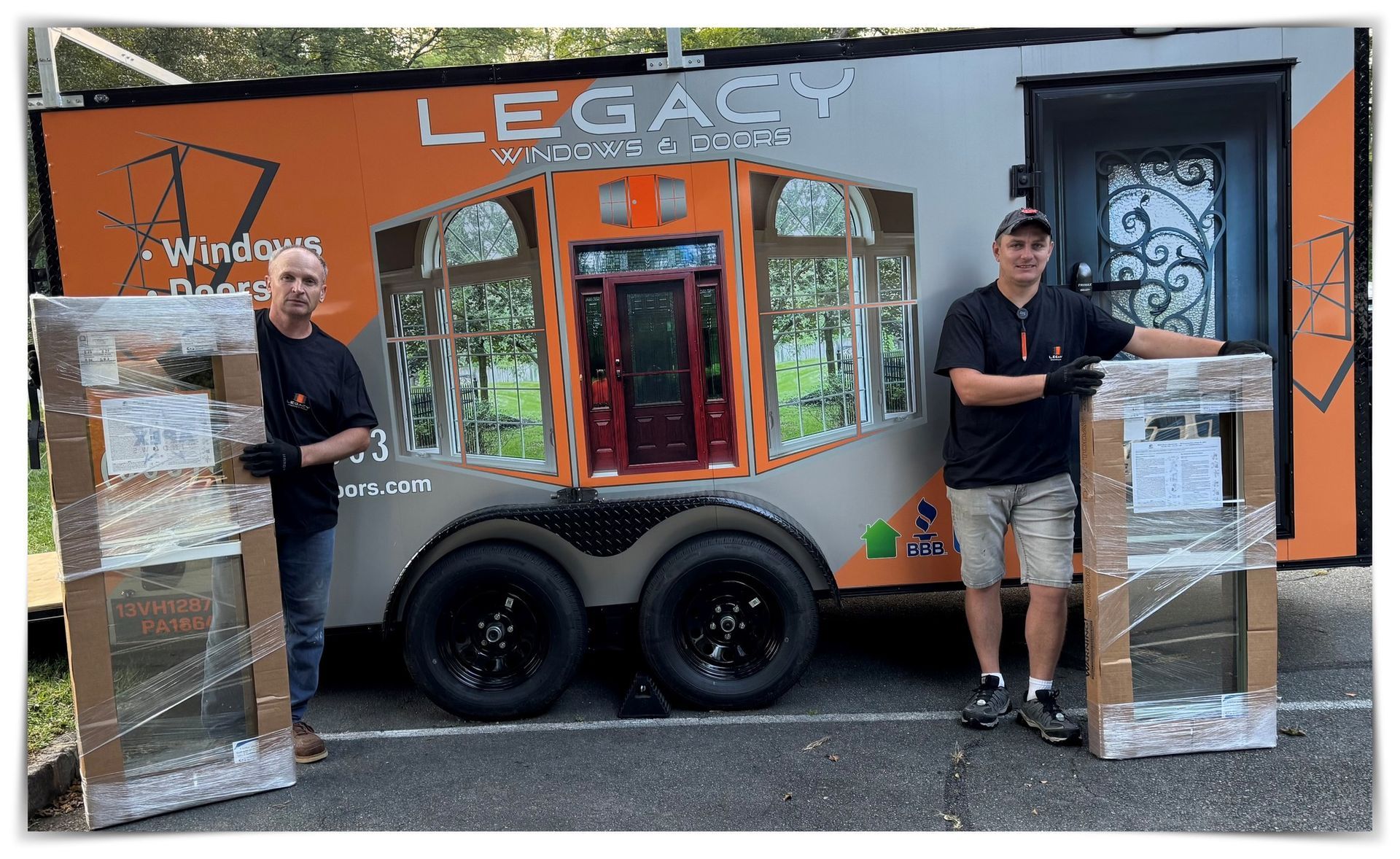 Two men stand by a trailer, holding packaged windows. The trailer has a logo and a door display.