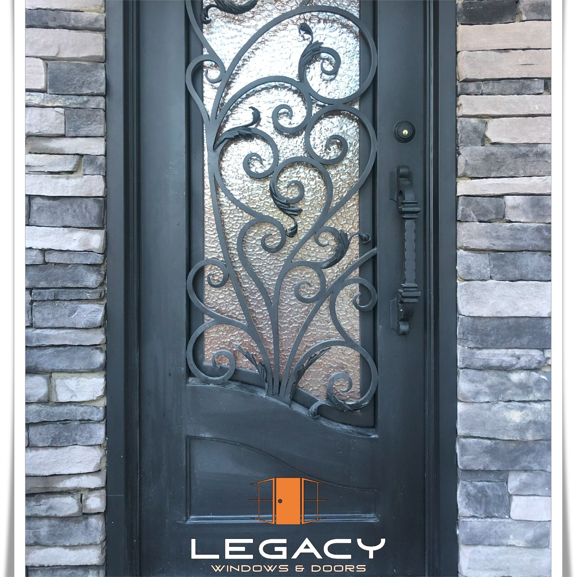 Black metal front door with decorative scrollwork and frosted glass panel, set in a stone wall.