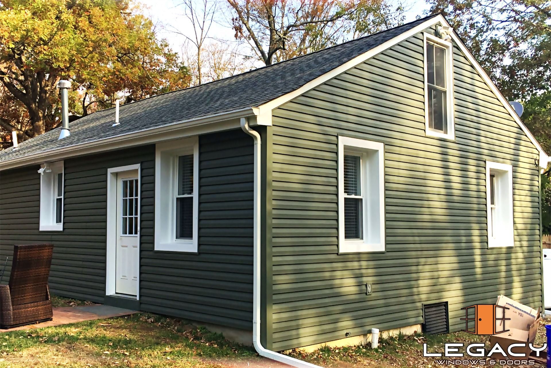 Green-sided house with white trim, windows, and door. Autumn trees in the background.