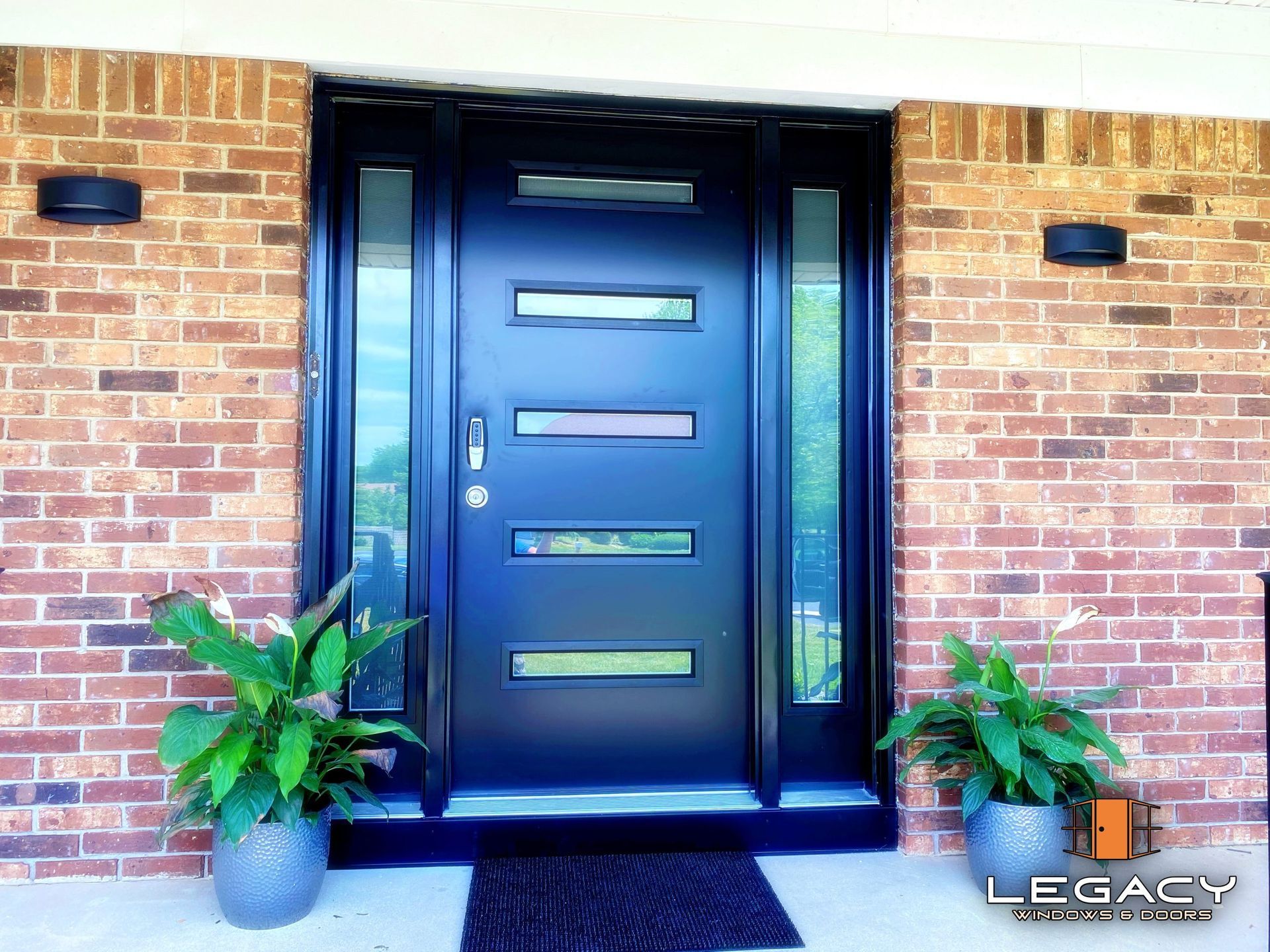 Black front door with sidelights, brick exterior, two potted plants.