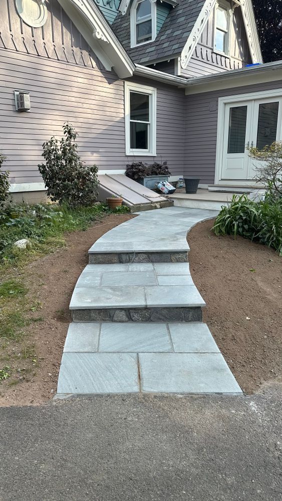 Stone steps leading to a light purple house entrance. A winding walkway with plants on either side is in the foreground.