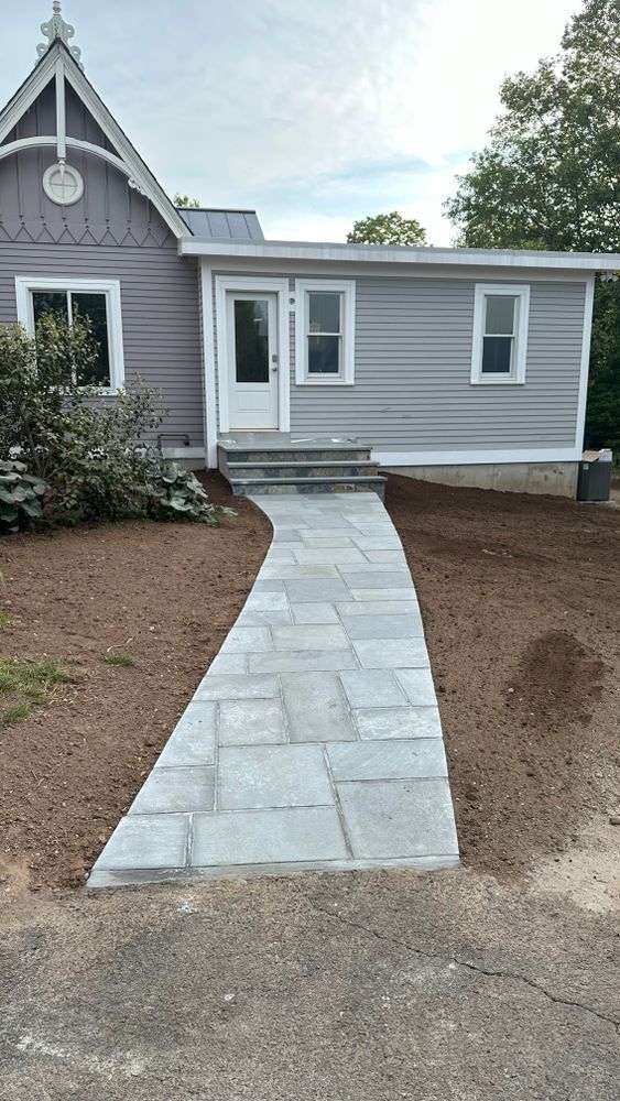 A gray stone pathway leads to a light gray house with white trim and a front door. Brown dirt borders the path.
