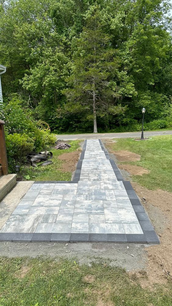 Paver walkway leading from a porch to a driveway, bordered by dark gray pavers. Lush green trees are in the background.