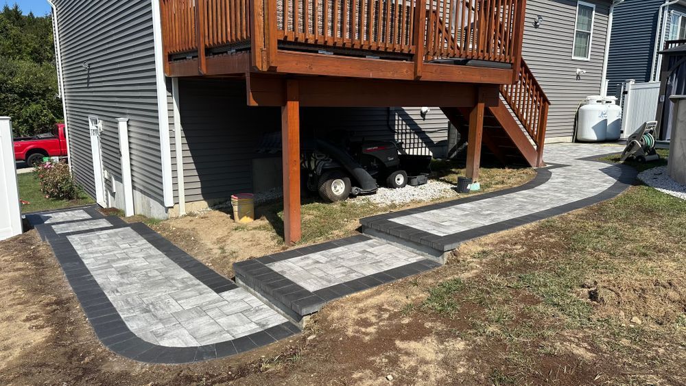 A paved walkway with steps leads under a wooden deck. The path is gray with a black border, surrounded by brown grass.