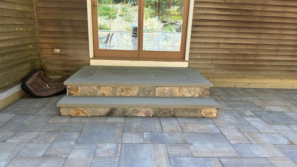 Stone steps leading to a doorway on a wooden building with a stone patio in the foreground; a dog bed rests on the steps.