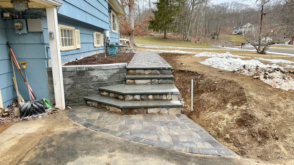 Stone steps and walkway leading up to a blue house. A section of the yard is dug up, with snow piles in the background.