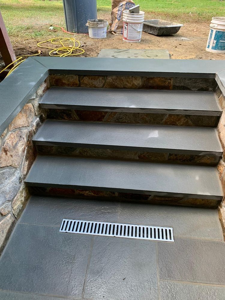 Stone steps with a drainage grate at the bottom. The steps are gray and lead up, set against a natural stone wall.