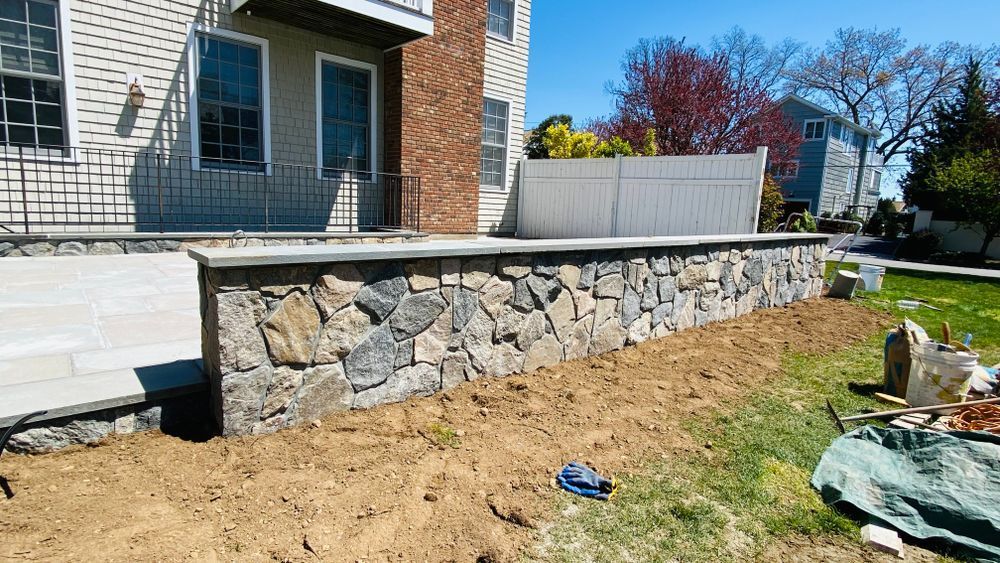 Stone retaining wall with gray cap, fronting a house with a brick chimney. Green grass and blue sky.